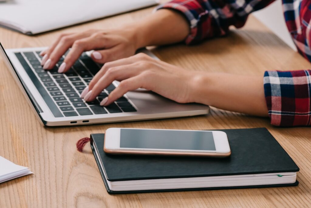 partial view of woman typing on laptop at tabletop with smartphone and notebook